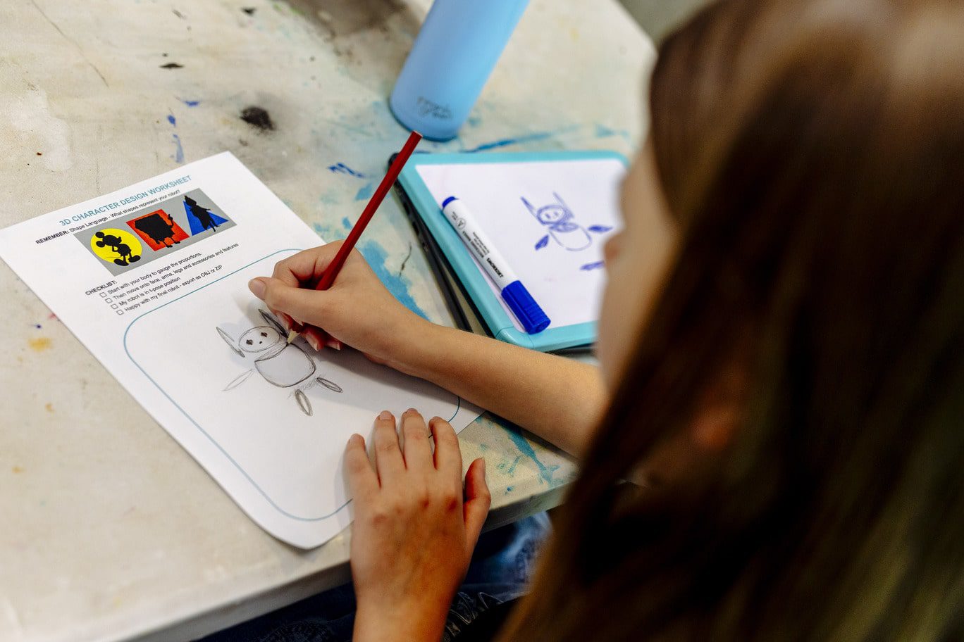 Blurred in the right-hand side of the foreground is a student with long straight brown hair. The student drawing a rounded geometric creature within a blue outlined rounded edge square on an A4 worksheet. The worksheet includes the blue capitalised title '3D CHARACTER DESIGN WORKSHEET' and includes simple imagery and a checklist. In the background is a paint stained light-grey table with a whiteboard to the right which features the same drawing of the rounded geometric creature in dark blue whiteboard marker.