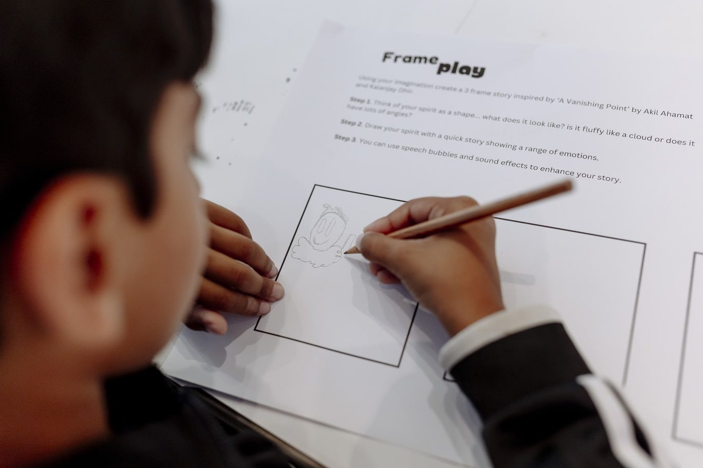 A young boy is draws a simple happy face atop a cloud within a comic stip style box on a white A4 paper entitled 'Frame play'. The photo is taken from behind the child, whose face is blurred on the left hand side of the image, and his right hand, which is drawing with a wooden graphite pencil, is central in view.