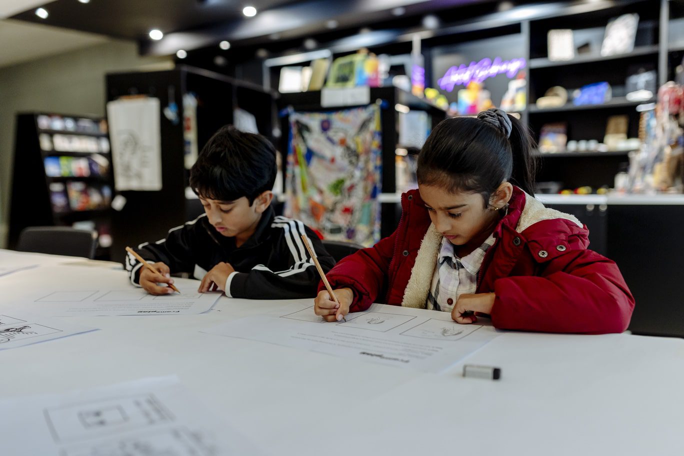 A young boy and girl are pictured drawing images within the three comic strip boxes on A4 worksheets entitled 'Frame play'. The children are looking down at their drawings, both sitting side-by-side at a white table, scattered with other completed worksheets. Blurred in the background is the Campbelltown Arts Centre's 'Artist Exchange' shop with black cabinets and filled with colourful wares.