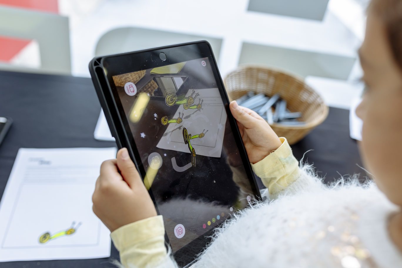 A young girl is pictured from the side as she holds and iPad in front of a table with a a basket of pens and an A4 worksheet picturing a colourful hand-drawn snail. The iPad screen pictures an AR experience of five of the same hand-drawn snails moving around the screen in 3D, in different angles and orientations.