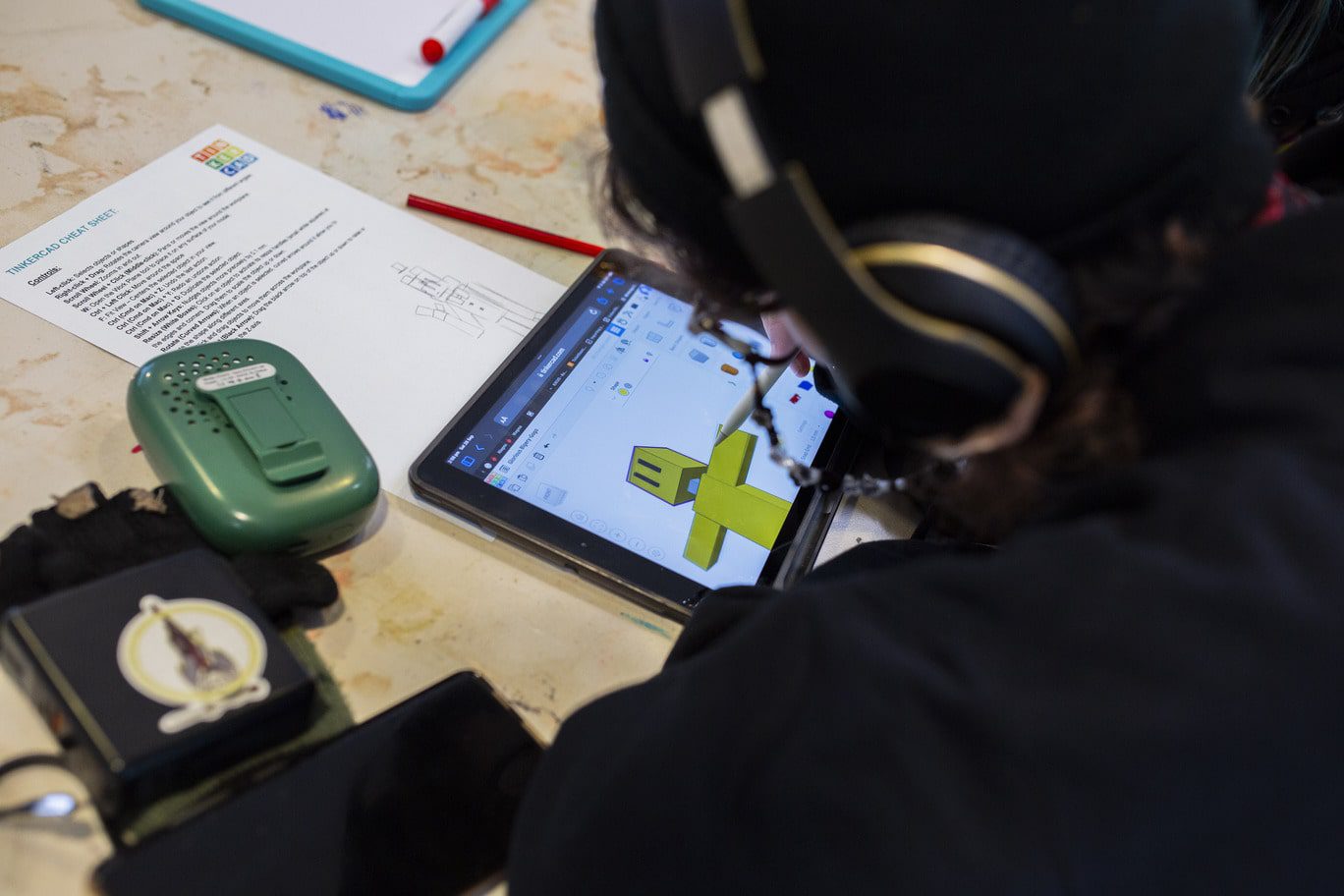 Blurred in the right-hand side of the foreground is a student with a black shirt, glasses and headphones. The student is using an Apple Pencil to draw an image of a geometric yellow robot on an iPad's drawing program. Scattred on the paint stained light-grey table are various objects including a green electronic device, a phone, a pencil, whiteboard and A4 worksheet entitled ' TINKERCARD CHEAT SHEET'.