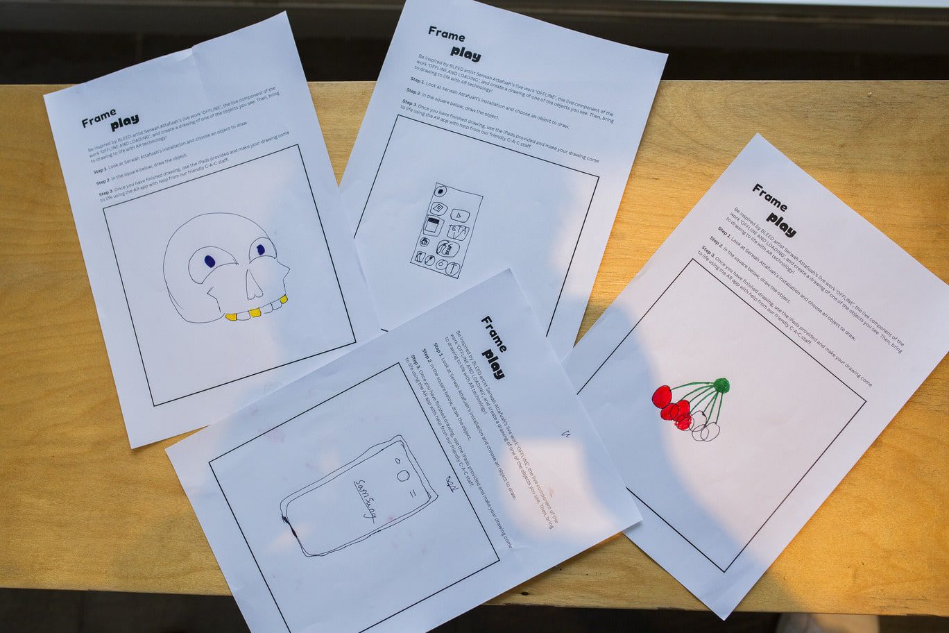 Scattered on a wooden bench are four overlapping portrait oriented A4 white worksheets. Each worksheet includes the title 'Frame play' with step-by-step instructions for the activity, and each includes a large black-outlined box which fills the page. Each A4 sheet features a unique hand-drawn image within the box. The sheet on the top left pictures a happy skull with three yellow teeth, the central top sheet pictures a black pen drawing of smart phone screen, the top right sheet pictures a bunch of red cherries with green stems and the bottom sheet features a simple drawing of a Samsung phone.