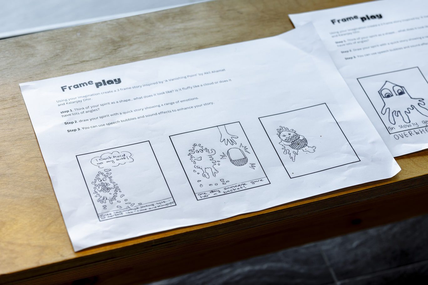 Two A4 white paper worksheets sit atop a wooden bench. Both in landsape orientation and entitled 'Frame play', the worksheets include step-by-step instructons and three boxes for comic book style drawings. Within the three boxes of the first worksheet are images drawn with a black pen which feature a character who uses a basket to help hold all it's 